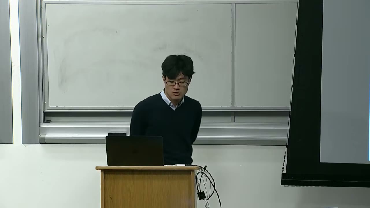 A speaker stands at a podium with a laptop, presenting in a lecture hall.