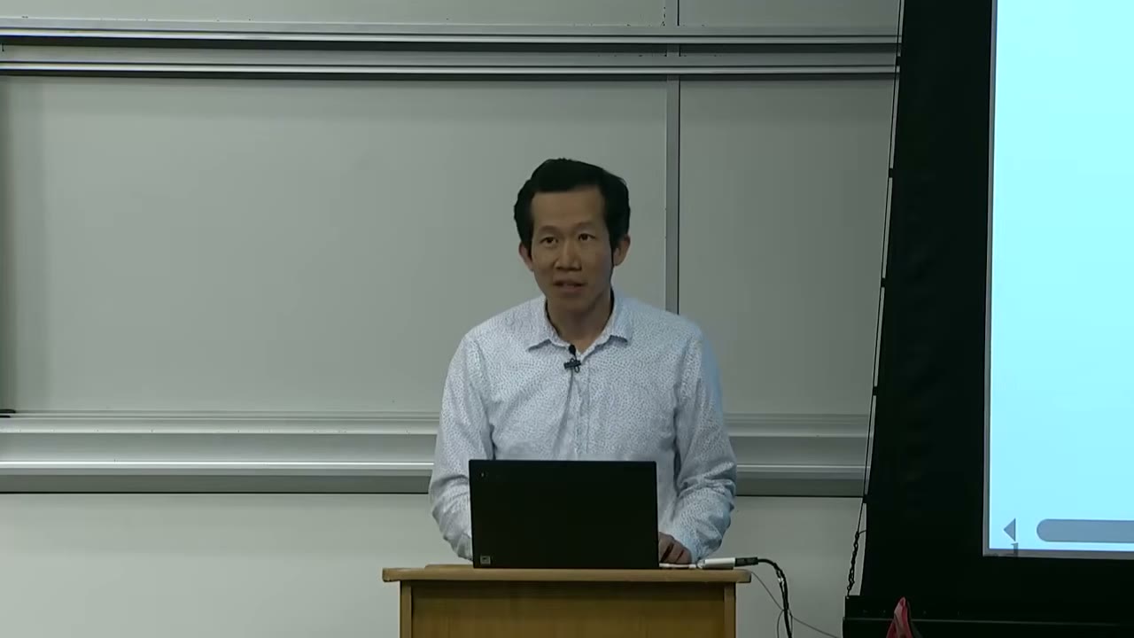 The speaker is standing at a podium with a laptop, facing the audience.