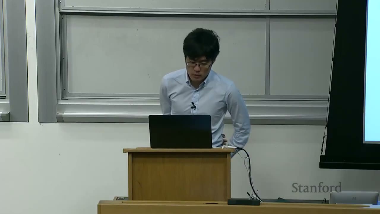 A male lecturer stands at a podium, looking at his laptop.