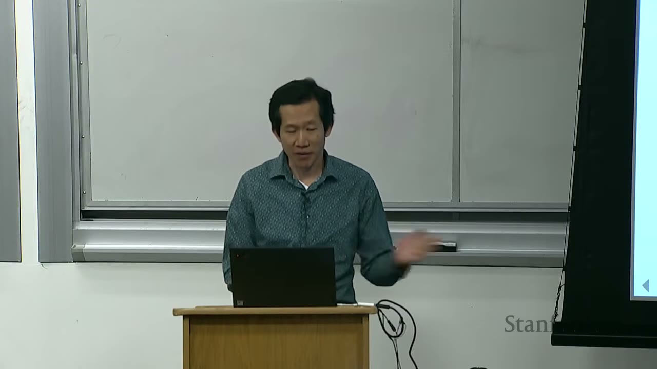 A lecturer stands behind a podium with a laptop, gesturing while speaking.