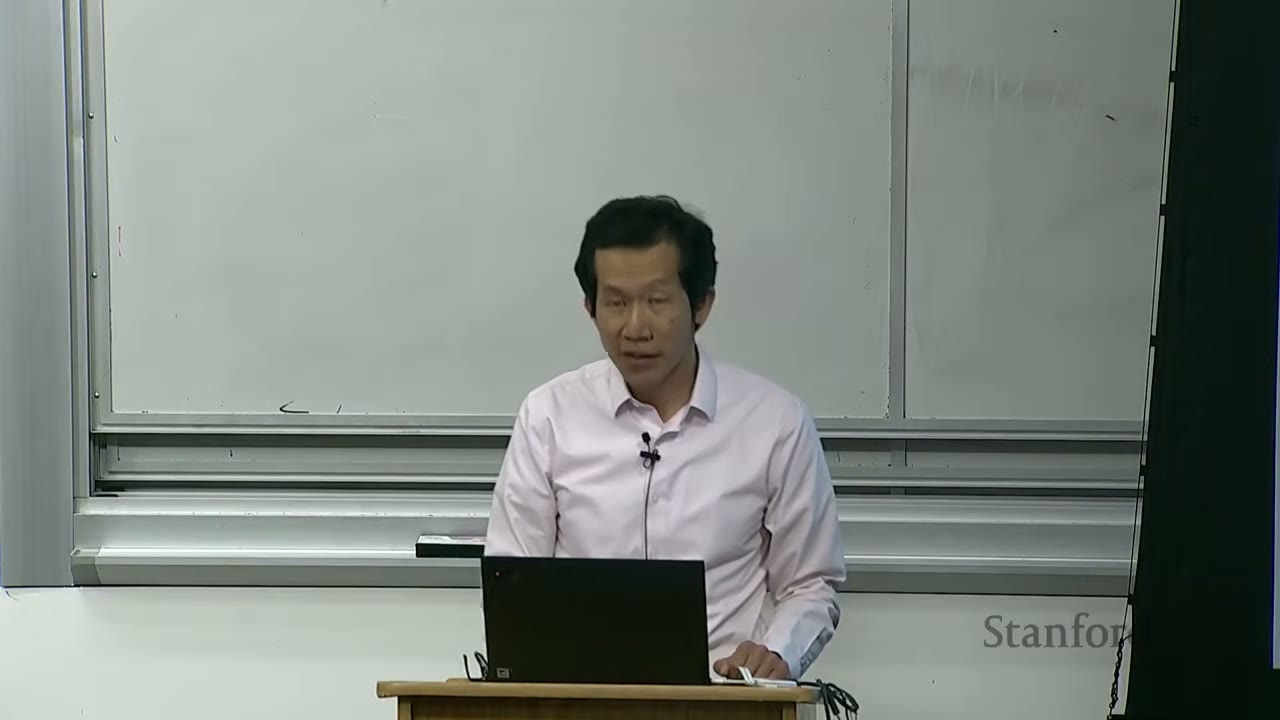 A speaker stands in front of a whiteboard, presenting a lecture with a laptop.